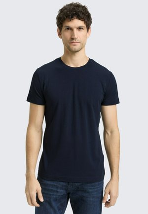 Mann mit lockigem dunklem Haar, der ein schlichtes schwarzes kurzärmeliges T-Shirt und dunkelblaue Jeans trägt, vor einem hellgrauen Hintergrund stehend.