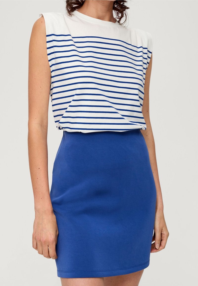 Femme portant un haut sans manches blanc avec des rayures horizontales bleues et une jupe ajustée bleu uni sur un fond uni.