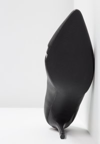 Chaussure à talon haut noire à bout pointu vue de la semelle, appuyée contre une surface blanche.