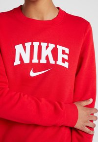 Röd sweatshirt i mjukt material, med stor vit "NIKE"-text och logotyp över bröstet, samt ribbade muddar och halsringning.