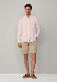 Helles rosa Leinenhemd mit Knöpfen und Kragen, kombiniert mit beigen Shorts und braunen Bootsschuhen. Einfaches Design, leichtes Material, lässiges Outfit.