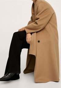 Manteau en laine couleur camel à coupe décontractée, grands revers et fermeture à un bouton. Associé à un pantalon noir évasé et des bottes noires élégantes.