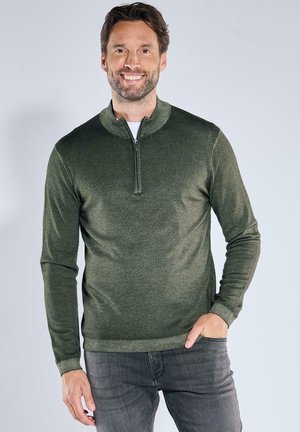 Dunkelgrüner Pullover mit Reißverschlusskragen, gestrickter Textur und langen Ärmeln. Trägt man über einem weißen Hemd, kombiniert mit grauen Jeans.