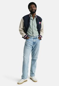 Giacca varsity blu navy e beige con polsini a coste a righe, camicia a righe verde, jeans azzurri chiari e sneaker bianche con dettagli grigi.