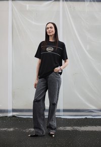 Černé tričko s potiskem a logem, spárované s vysokým pasem, roztrhanými šedými džínami a černými botami s kovovými doplňky. Postava stojí na neutrálním pozadí.