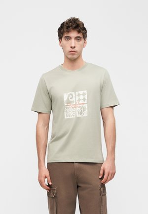 Jeune homme aux cheveux courts et bouclés, portant un t-shirt graphique vert clair et un pantalon cargo marron, debout devant un fond blanc uni.