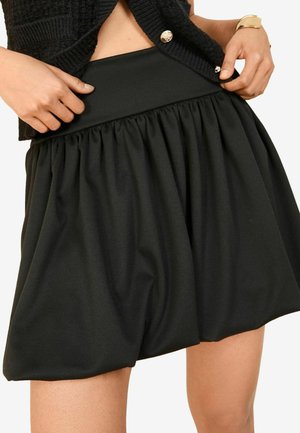 Kvinde iført en sort rynket mininederdel og en sort strikket cardigan med guldknap og armbånd, hænder justerer linningen.