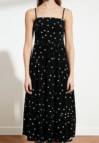 Kobieta w czarnej, bezrękawnikowej sukience midi z białymi wzorami gwiazd i półksiężyca na tle gładkiej białej ściany.