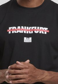 Zwarte katoenen t-shirt met witte en rode grafische tekst "FRANKFURT RU(I)NNING THE NEIGHBOURHOOD" en het logo "WEEKEND OFFENDER". Eenvoudige ronde halslijn.