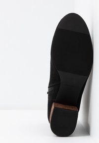 Botte en daim noir avec fermeture éclair latérale, semelle en caoutchouc et talon en bois empilé, positionnée droite contre une surface d'angle blanche.