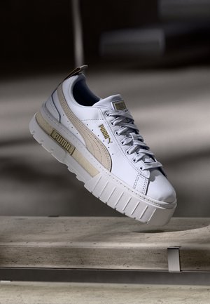 Hvid Puma-sneaker med beige logostribe og chunky sål, svævende i luften over en betonbænk i en dæmpet belysning.