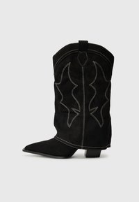 Botte de cowboy en suede noir avec un bout pointu, un petit talon et des coutures décoratives blanches, présentant un motif en forme de flamme sur la tige.