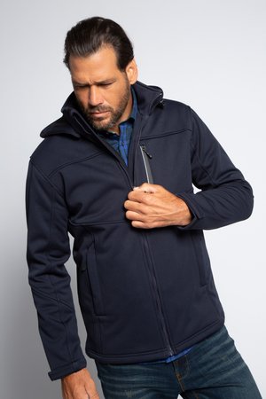 Uomo con giacca blu navy che apre la tasca al petto, vestito con camicia a quadri blu e jeans, in piedi davanti a uno sfondo chiaro e semplice.