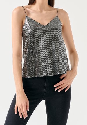 Mujer con una camiseta sin mangas de lentejuelas plateadas con tirantes finos y pantalones negros ajustados, posando con una mano en la cadera y la otra relajada.
