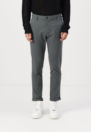 Graue Baumwollmischungshosen mit flachem Bund, schmal zulaufenden Beinen und umgeschlagenen Säumen, kombiniert mit weißen Turnschuhen und einem strukturierten schwarzen Pullover.