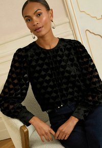 Melna blūze ar sirds rakstu, puff piedurknēm un apaļo dekoltē. Audumam ir neliela spīduma pakāpe un tam ir krokas priekšpusē.