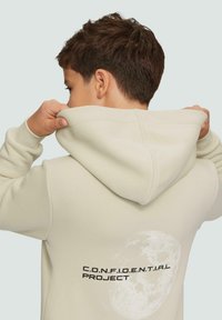 Lichtbeige hoodie met een capuchon met trekkoorden, voorzien van een maanafbeelding en de tekst "C.O.N.F.I.D.E.N.T.I.A.L PROJECT" op de achterkant. Gladde textuur.