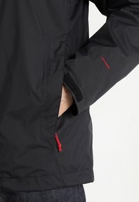 Manche de veste noire avec poignet Velcro réglable et main dans la poche latérale avec tirette de fermeture éclair rouge et étiquette "DRYVENT" sur la manche.