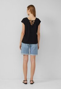 Vrouw in een zwart T-shirt met korte mouwen en een strikdetail op de achterkant, lichtblauwe denimshorts met omgerolde zoom en zwarte sandalen, staand tegen een plain muur.
