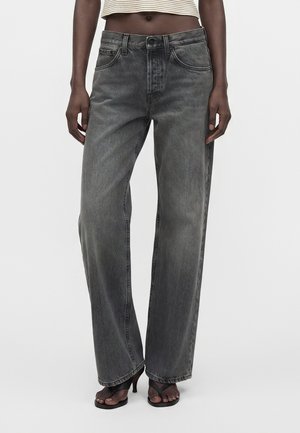 Person iført højtaljede, løstsiddende, falmede sorte jeans med en cropped stribet top og sorte hælede sandaler, stående mod en ensfarvet baggrund.