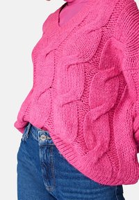 Personne portant un pull tricoté à gros câbles d'un rose vif par-dessus un col roulé fuchsia, associé à un jean en denim bleu taille haute sur fond blanc.