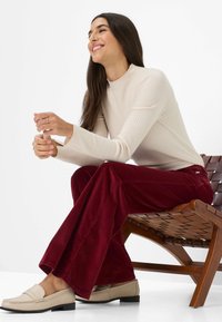 Beige gerippter Langarmshirt, bordeauxfarbene Cordhosen mit Schlag und hellbeige Loafers. Sitzt auf einem Holzlattenstuhl.