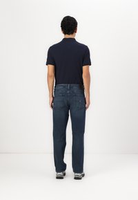 Polo shirt bleu foncé, jean droit bleu avec poches visibles et baskets noires, vu de dos sur un fond blanc.