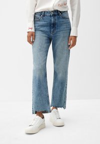 Jeans de denim azul acampanados con dobladillo sin terminar, llevados con zapatillas blancas. Cuenta con bolsillos frontales y un lavado medio, destacando áreas desgastadas.