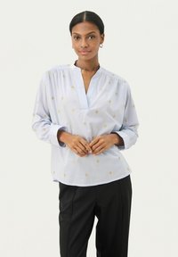 Blusa azzurro chiaro a maniche lunghe con scollatura a V profonda, decorata con motivi a diamante dorati. Tessuto morbido con leggero arricciamento a soffietto sulle spalle. Pantaloni neri.