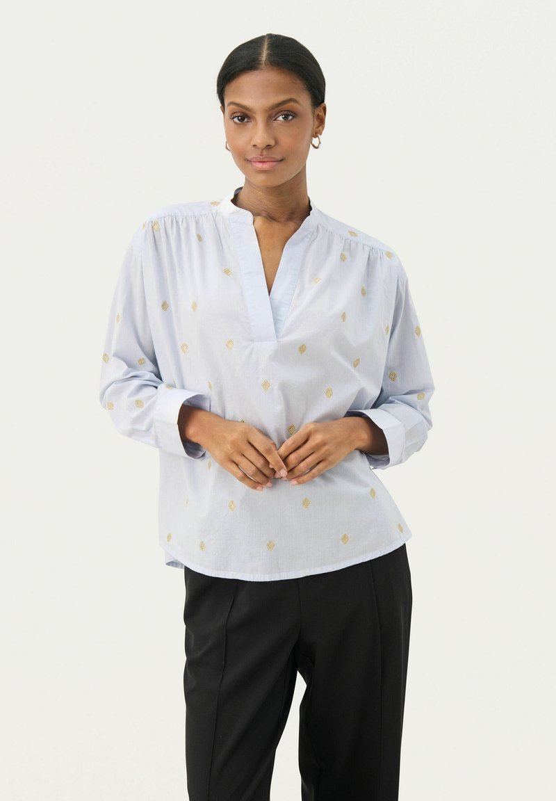 Blusa azzurro chiaro a maniche lunghe con scollatura a V profonda, decorata con motivi a diamante dorati. Tessuto morbido con leggero arricciamento a soffietto sulle spalle. Pantaloni neri.