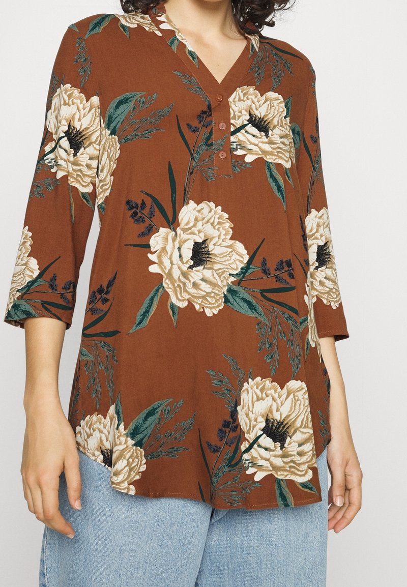 Brązowa bluzka w kwiaty z dużymi kremowymi i zielonymi kwiatami, rękawami do łokci, dekoltem w serek oraz detalami w postaci guzików z przodu.