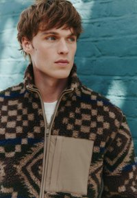 Fleece jacket in brown, green, and blue geometric patterns with a tan pocket. Features a high collar and zip closure. Soft texture.