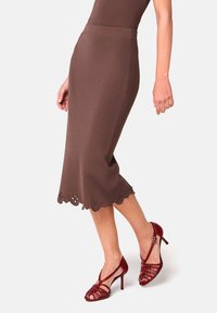 Jupe crayon marron en tissu extensible, dotée d'un ourlet festonné avec des découpes florales. Associée à des chaussures à talons hauts rouges à lanières.