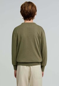 Olivgrüner Strickpullover mit Rundhalsausschnitt, überschnittenen Schultern und gerippten Bündchen. Kombiniert mit hellbeigen Hosen mit einer Gesäßtasche.