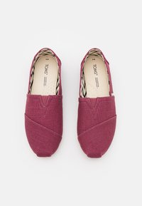 Two burgundy canvas slip-on shoes featuring a textured surface, V-shaped stitching detail, and a patterned interior lining.