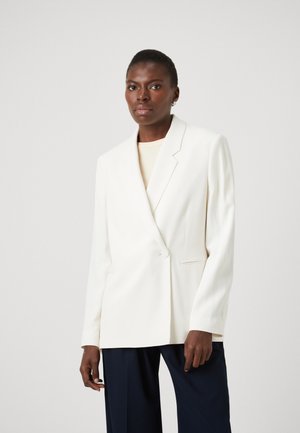 White tailored blazer with peak lapels, single button closure, and two front pockets. Worn over a light top with wide-leg navy trousers.