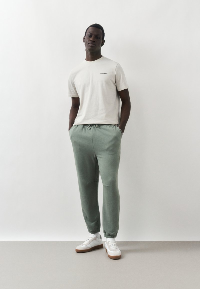 Un jeune homme est détendu, appuyé contre un mur blanc, portant un t-shirt Calvin Klein écru, un jogging vert clair et des baskets blanches avec des semelles marron.