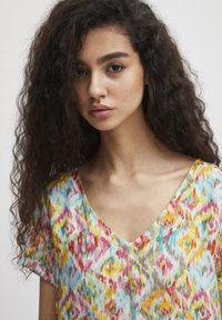 Blouse multicolore avec un décolleté en V, des manches courtes et un tissu léger et texturé arborant un motif abstrait semblable à de l'aquarelle.