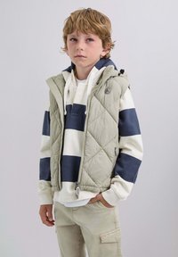 Niño con cabello castaño claro, vistiendo una sudadera a rayas blanca y marino, un chaleco ligero acolchado y pantalones cargo beige, de pie con una mano en el bolsillo.