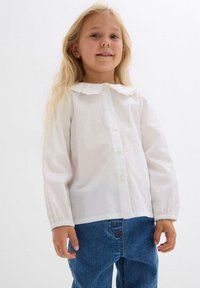 Camicia bianca a maniche lunghe con colletto arricciato, dotata di chiusura frontale con bottoni. Abbinata a jeans in denim blu. Texture del tessuto liscia.