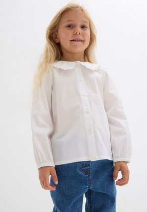 Camicia bianca a maniche lunghe con colletto arricciato, dotata di chiusura frontale con bottoni. Abbinata a jeans in denim blu. Texture del tessuto liscia.