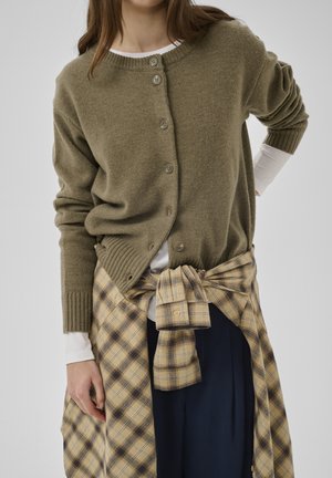 Frau trägt einen olivgrünen, geknöpften Cardigan über einem weißen Langarmshirt mit einem beige karierten Hemd, das um die Taille einer marineblauen Hose gebunden ist.