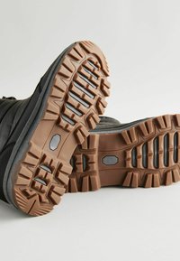 Chaussures noires avec des semelles en caoutchouc robuste dotées de profondes rainures, de détails de surface texturée et d'une semelle extérieure marron contrastante pour une meilleure adhérence et durabilité.