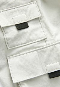 Tissu gris clair avec une texture lisse comportant deux poches à rabat fixées par des languettes noires. Les coutures sont visibles le long des bords.