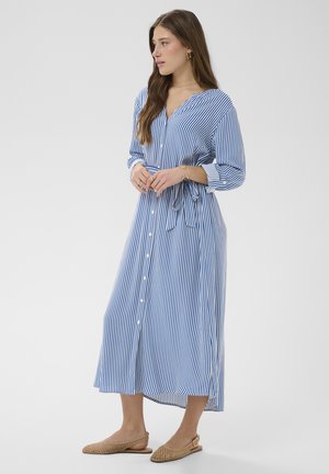 Kobieta ubrana w długą sukienkę typu koszulowego z pionowymi niebiesko-białymi paskami, z długimi rękawami, przewiązaną w talii, nosząca beżowe plecione sandały na płaskim obcasie z paskiem wokół pięty.
