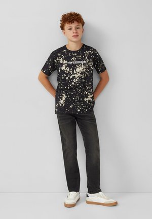 Zwarte katoenen t-shirt met een wit gespikkeld patroon, ronde hals, korte mouwen; gecombineerd met donkere jeans en witte sneakers met een bruine zool.