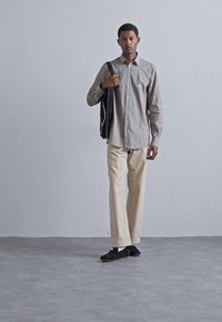 Model îmbrăcat cu o cămașă cu dungi subtiri de culoare maronie deschisă, pantaloni bej și pantofi slip-on negri, purtând o geantă tote neagră. Fundal simplu.