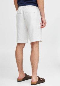 Hvite linshorts med elastisk midje, to baklommer og avslappet passform, kombinert med brune sandaler på en lys bakgrunn.