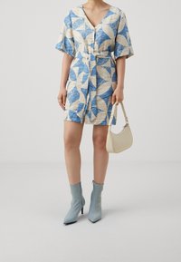 Robe à manches courtes avec un motif géométrique bleu et beige, cintrée à la taille. Associée à des bottines bleu clair et un sac à main crème.