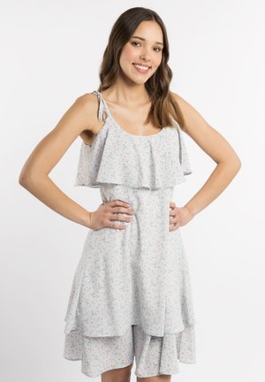 Junge Frau lächelt, Hände in den Hüften, trägt ein hellblaues ärmelloses Kleid mit Blumenmuster und Rüschenschichten vor weißem Hintergrund.